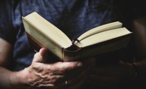 eine ältere Hand hält ein aufgeschlagenes Buch, in dem die Person offenbar liest